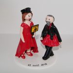 figurine de mariage avec mariée en robe rouge, tenant une pancarte bus scolaire, et le marié en costume avec tutu rouge et chaussons de danse
